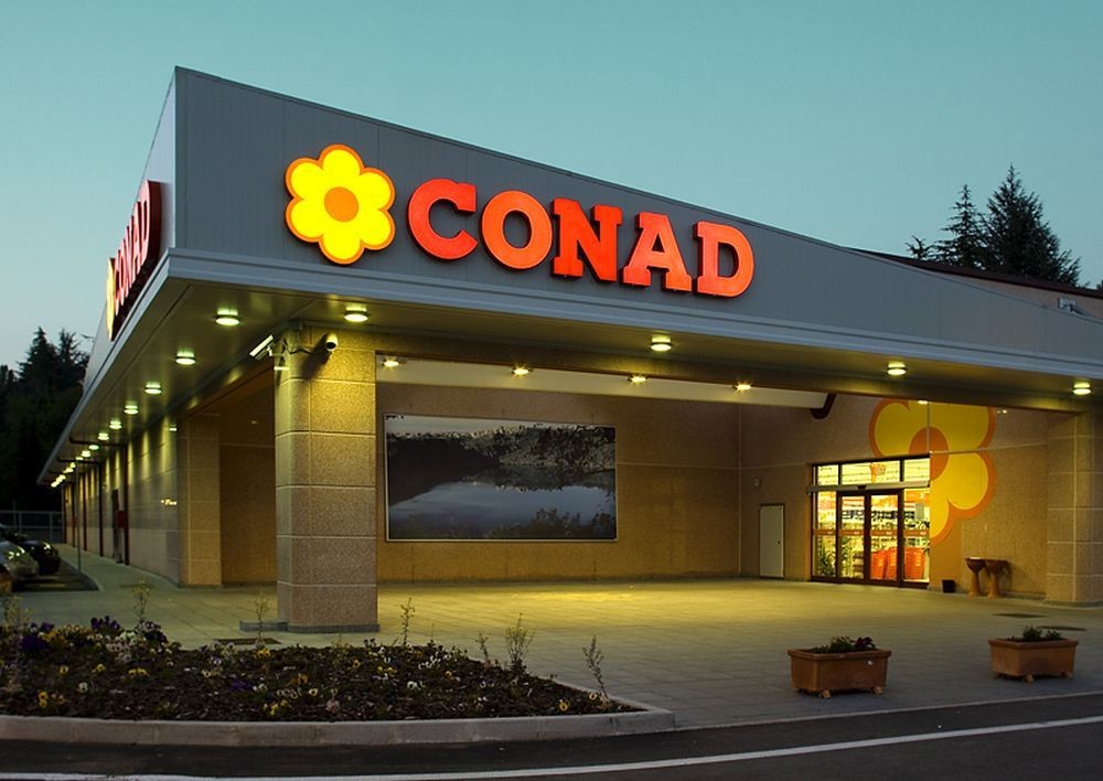 Exterior view of a Conad supermarket building with illuminated signage and a flower logo.